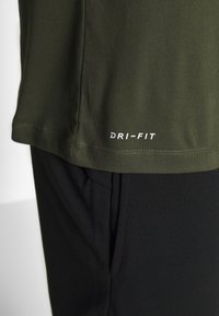 Camiseta Dri-FIT color verde oliva con una textura suave, corte ligeramente suelto y un logo blanco en el dobladillo, combinada con pantalones deportivos negros.