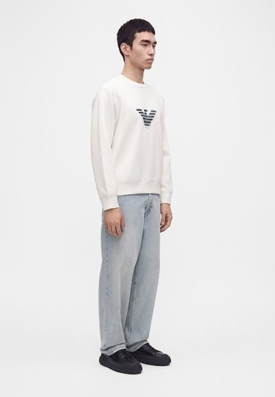 Homme portant un sweat-shirt blanc avec un logo noir, un jean bleu clair à jambes larges et des baskets noires, debout devant un fond blanc uni.