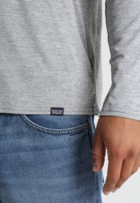 Maglietta grigia a maniche lunghe con tessuto testurizzato, con un'etichetta sul bordo recante il logo Patagonia, abbinata a jeans in denim blu.