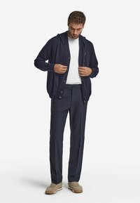 Mörkblå cardigan med huva, knäppning fram, lagd över en vit t-shirt, matchad med mörkblå byxor och beige sneakers.