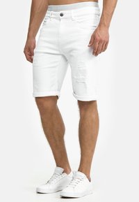 Witte denim shorts met gescheurde accenten, opgerolde mouwen en een lichtgrijze tailleband met logo-tekst; gecombineerd met witte sneakers.