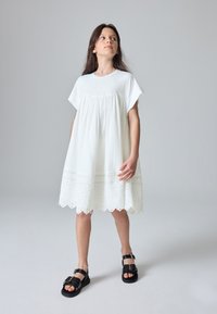 Hvid bomuldskjole med løs pasform, korte ærmer, rynket overdel og blonder ved kanten. Kombineret med sorte sandaler med spænder.