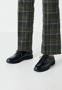 Scarpe eleganti nere in pelle verniciata con punta arrotondata, design con lacci e superficie testurizzata, abbinate a pantaloni a quadri verdi e neri.
