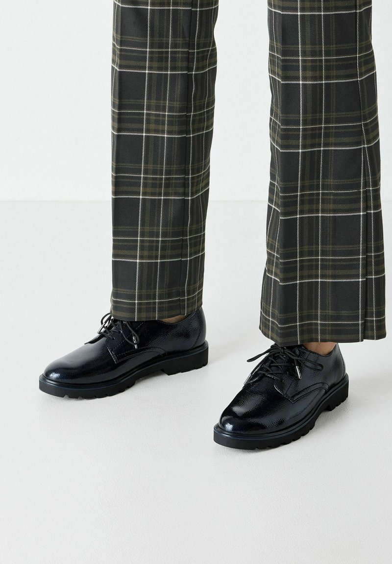 Scarpe eleganti nere in pelle verniciata con punta arrotondata, design con lacci e superficie testurizzata, abbinate a pantaloni a quadri verdi e neri.