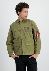 Chaqueta ligera de color verde oliva con múltiples bolsillos, botones a presión y tira de cierre rojo. Presenta texto impreso y parches en la parte frontal.