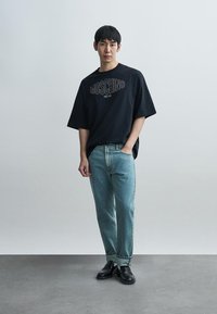 Μαύρο υπερμεγέθες μπλουζάκι με το κείμενο «MOSCHINO», ανοιχτό μπλε τζιν παντελόνια και μαύρα παπούτσια, όλα τοποθετημένα σε ουδέτερο φόντο.