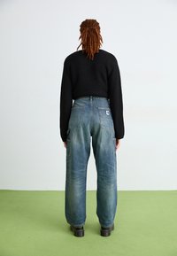 Maglione nero a coste, jeans blu sbiaditi a vestibilità ampia con molteplici tasche e una piccola etichetta con logo, abbinati a calzature nere.