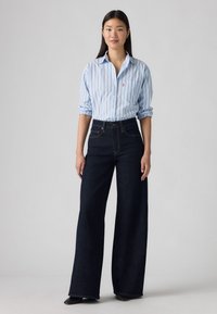 Camisa azul claro com riscas verticais, com botões e mangas arregaçadas, combinada com jeans de perna larga azul escura de cintura alta e sapatos pretos.