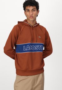 Ruda gaubta sweatshirt su mėlynu juostu, ant kurios baltais rašmenimis užrašyta "LACOSTE". Pagaminta iš lygaus audinio, su laisvu siluetu ir gumbuoto apvadu.