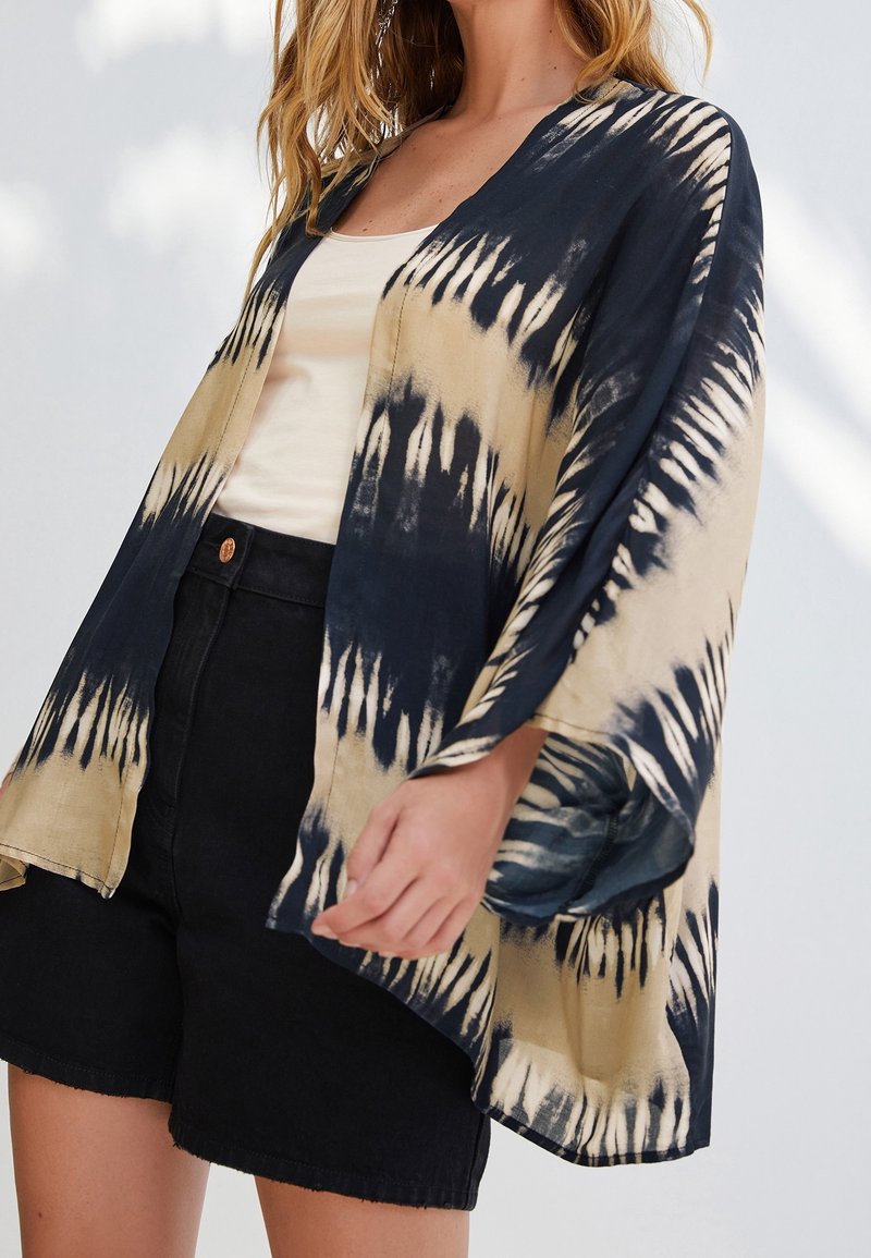 Tie-dye kimono met een beige en marineblauw patroon; lichtgewicht stof, losse pasvorm, open voorkant en wijde mouwen, gecombineerd met zwarte shorts.