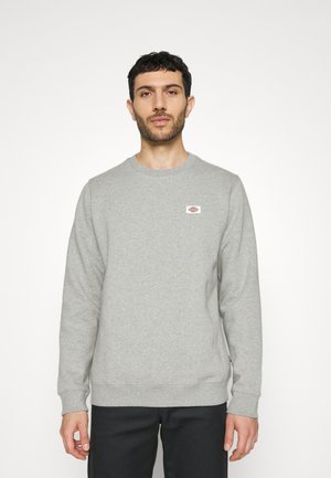 Sudadera gris de cuello redondo hecha de una tela suave, con un pequeño parche de logo en el pecho, puños y dobladillo acanalados.
