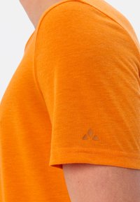 Korte mouwen oranje T-shirt met een gestructureerde stof; heeft een klein bruin logo op de mouw; het stiksel is netjes en gelijkmatig.