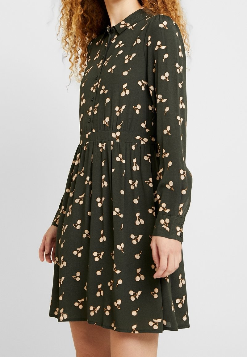 Femme portant une robe boutonnée à manches longues de couleur vert foncé avec un motif de cerises beige et une jupe plissée, debout devant un fond uni.