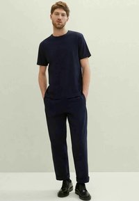 Young man standing with hands in pockets, wearing a navy blue short-sleeve shirt, matching pants, and black lace-up shoes.
