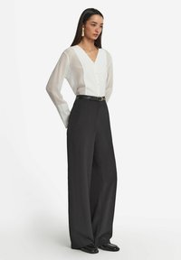 Blusa trasparente bianca con scollo a V e bottoni, abbinata a pantaloni neri a vita alta con gamba larga e una cintura nera. Tessuto morbido e vestibilità sartoriale.