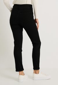 Jean en denim noir à coupe ajustée, taille mi-haute, poches arrière et bords retroussés, associé à des baskets blanches. Texture lisse et design classique.