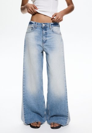 Un jean large bleu clair présente un lavage délavé, un design à cinq poches et une fermeture à bouton en métal. Le tissu affiche une texture lisse avec un minimum d'usure.
