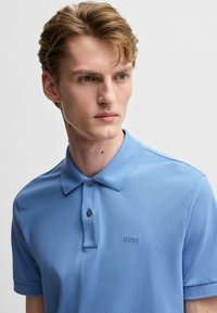 Ljusblå pikétröja i texturerad tyg, med klassisk krage, två knappar och en diskret broderad logotyp på bröstet.