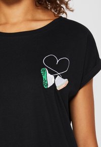 Svart t-shirt med färgglada broderade sneakers och en hjärtformad kontur. Korta ärmar och en avslappnad halsringning.