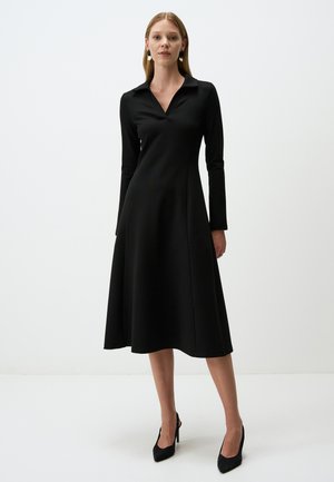 Robe noire à manches longues avec un col, silhouette évasée et tissu doux. Assortie à des chaussures à talons hauts noires.