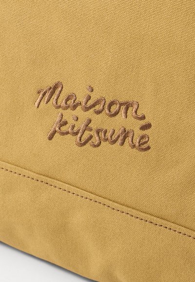 Sinatra dzeltena kanvas soma ar izšūtu "maison kitsuné" logotipu brūnā krāsā. Strukturēta virsma ar redzamām šuvju detaļām.