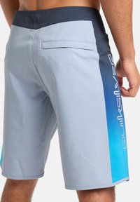 Sivé plážové šortky s tmavomodrým pásom, s gradientným modrým akcentom a bielym logom na boku, hladká tkanina, voľný strih.