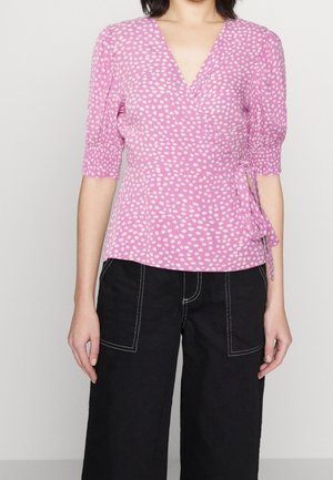 Femme portant un chemisier cache-cœur rose à manches courtes avec des pois blancs et un pantalon noir avec de grandes poches cousues en blanc.