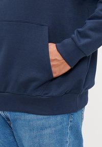 Marineblå hættetrøje med kengurulomme samt ribbede manchetter og kant. Blødt stof og afslappet pasform, kombineret med blå jeans.