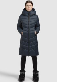 Tengerész kék, kapucnis puffertömörítő kabát, chevron steppeléssel, cipzárral és oldalsó zsebekkel. Fekete leggings és csizmák kíséretében viselve.