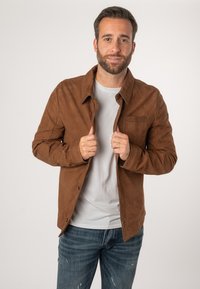 Bruine suede jas met een knoopsluiting aan de voorkant, twee borstzakken en een ontspannen pasvorm, gedragen over een lichtgrijs t-shirt en blauwe jeans.