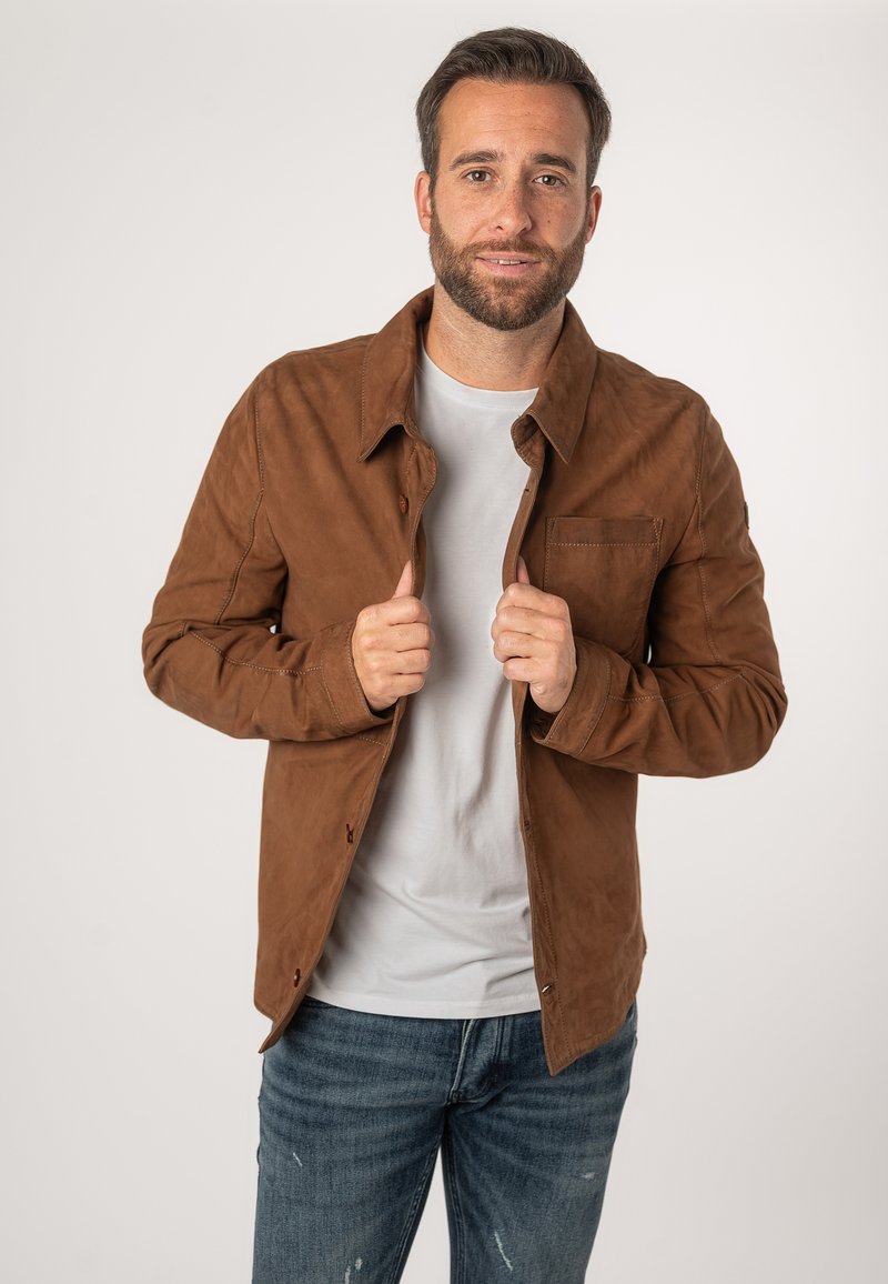 Bruine suede jas met een knoopsluiting aan de voorkant, twee borstzakken en een ontspannen pasvorm, gedragen over een lichtgrijs t-shirt en blauwe jeans.