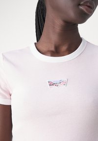 T-shirt rose clair avec un col blanc. Présente un patch logo coloré brodé sur la poitrine, fabriqué en coton doux.