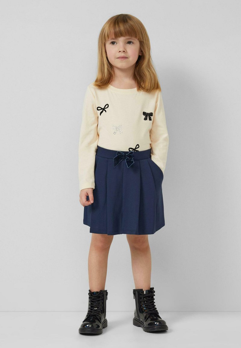 Kremowa bluzka z długim rękawem, ozdobiona czarną kokardą i kryształowymi akcentami, zestawiona z granatową plisowaną spódnicą i czarnymi, sznurowanymi botkami.