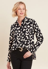 Camicia nera con motivo astratto bianco, maniche lunghe, colletto abbottonato, orlo curvo, abbinata a pantaloni neri e una cintura decorativa.