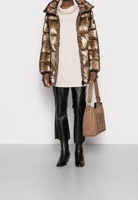 Mujer vestida con una chaqueta de plumas metálica bronce, vestido de suéter crema, pantalones de cuero negros cortos, botas negras hasta el tobillo, sosteniendo un bolso de ante beige.