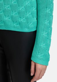 Türkisfarbener Strickpullover mit einem strukturierten Zopfmuster, mit einem gerippten Saum und Bündchen. Kombiniert mit schwarzen eng anliegenden Hosen.