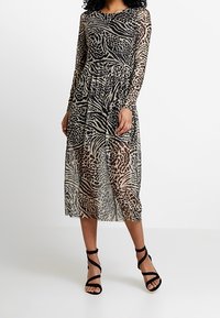 Genomskinlig klänning med långa ärmar som har ett leopardmönster i beige och svart, med en insydd midja och en svallande midi-längd. Svarta remsandaler.