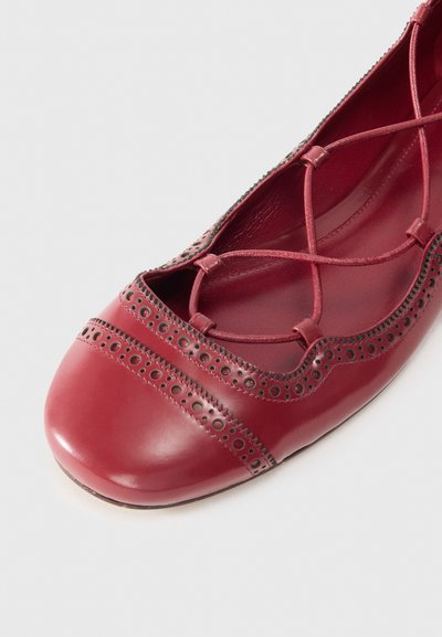 Red leather ballet flat with crisscross straps, perforated detailing around the opening, and a rounded toe. Smooth texture and polished finish.