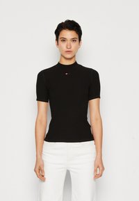 T-shirt noir ajusté à manches courtes, col montant, avec un logo rouge sur la poitrine. Texture lisse, tissu côtelé, porté avec un pantalon blanc.