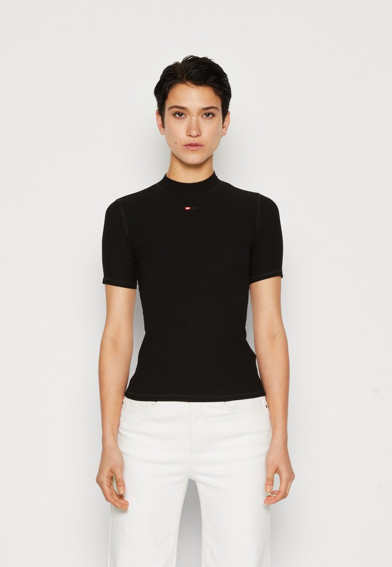T-shirt noir ajusté à manches courtes, col montant, avec un logo rouge sur la poitrine. Texture lisse, tissu côtelé, porté avec un pantalon blanc.