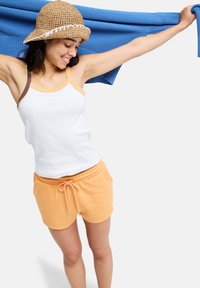 Valkoinen hihaton toppi, jossa oranssit ja ruskeat yksityiskohdat, yhdistettynä oransseihin narulla kiristettäviin shortseihin. Sininen neulospaita liehuu käsivarsilla.