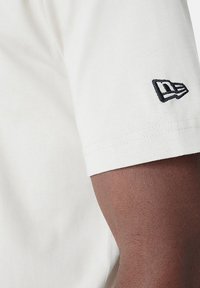 T-shirt à manches courtes, couleur beige clair, en coton, avec un petit logo noir brodé sur la manche. Texture lisse et design ajusté.