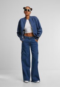 Džínová bunda so zipsom, široké cargo džínsy a biele crop top. Slnečné okuliare s hladkým rámom. Všetky položky v rôznych odtieňoch modrej.