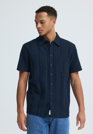 Jonge man met een marineblauw kortärmelig overhemd met knopen en een blauwe spijkerbroek, kijkt naar beneden en iets opzij tegen een effen achtergrond.