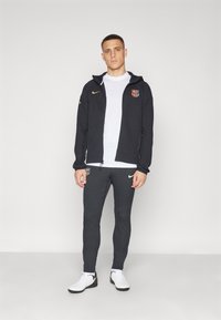 Felpa nera con zip e logo, t-shirt bianca, pantaloni grigio scuro texturizzati e scarpe da ginnastica bianche con accenti neri.