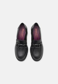 Mocassins noirs en cuir avec un accent chaîne, présentant une texture lisse et un bout arrondi. Semelle intérieure violette avec détail de marque.