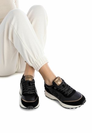 Zapatillas negras con materiales de ante y cuero, con un acento en bronce, sistema de cordones en la parte frontal y suela blanca. Combinadas con joggers de color claro.