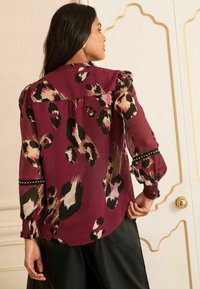 Bordeauxfarbene Bluse mit Leopardenmuster, durchsichtigen Materialien, aufgebauschten Ärmeln und gerüschten Manschetten. Verfügt über dekorative Akzentstreifen und einen abgerundeten Saum.