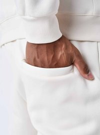 Sudadera blanca con puños de canalé, primer plano de una mano descansando en un bolsillo blanco; textura suave con detalles de costura mínimos.