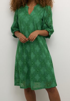 Kvinde iført en knælængde grøn blondekjole med 3/4-ærmer, stående foran en enkel lysvæg.